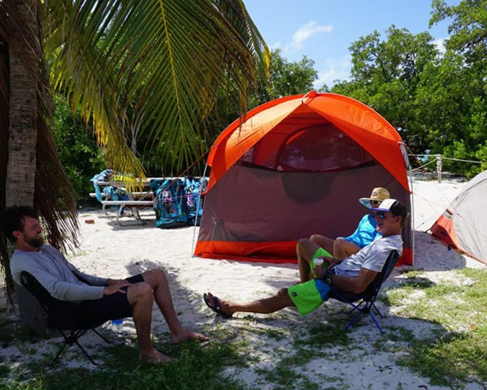 three people sit on camping chairs next to tents under palm trees