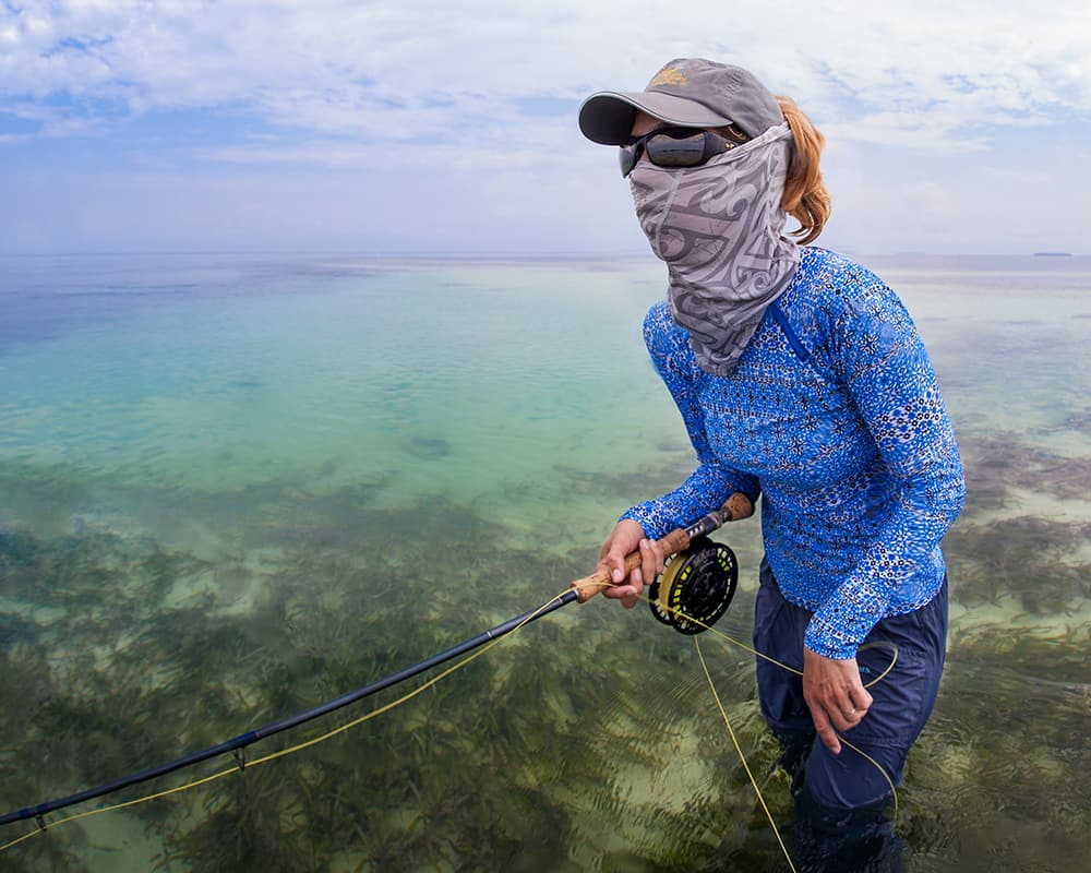 a person standing in shallow water holding a fishing rod and wearing a sun protective shirt and fishing buff