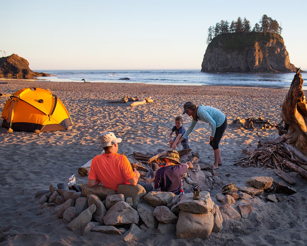 people gather by a fire pit on the beach with a tent in the foreground and the ocean in the background