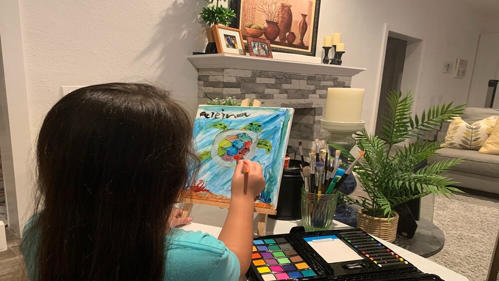 The back of a young girl in a blue shirt painting a sea turtle in her living room.