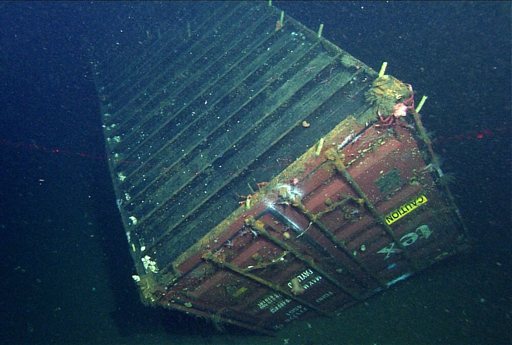 A metal shipping container lies upside down on the seafloor. White text is visible on one end.