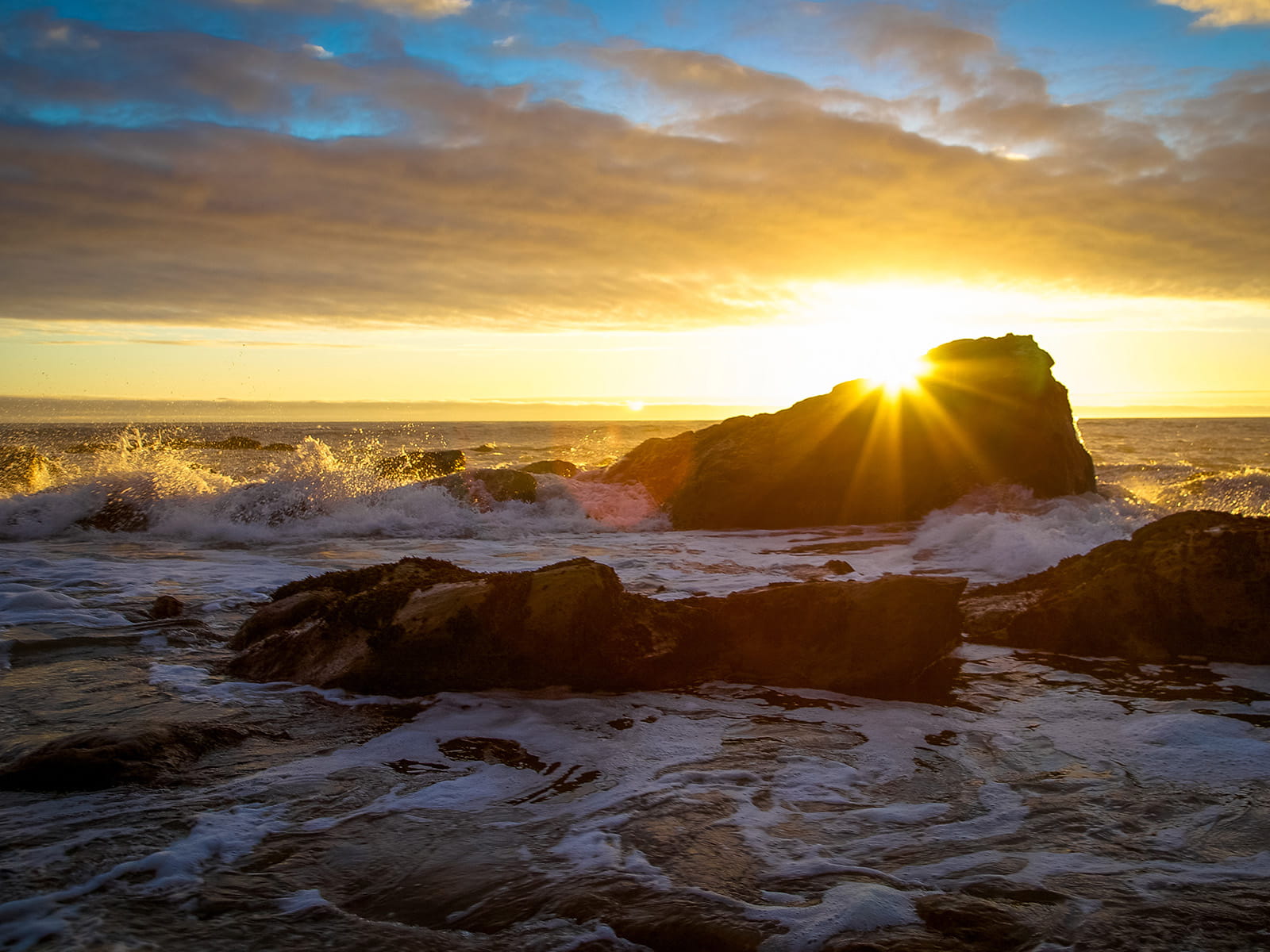 sunseting in the distance while waves crash over rocks along the beach