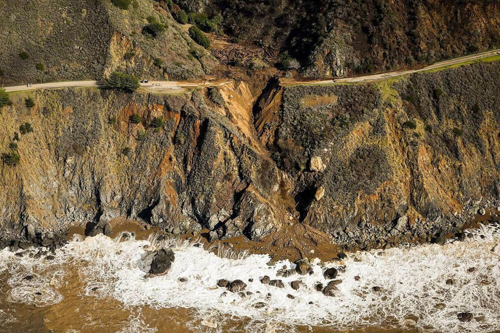 A road along a cliff on the shoreline is split at the side of a mudslide. Rocks and debris are visible at the shoreline below.