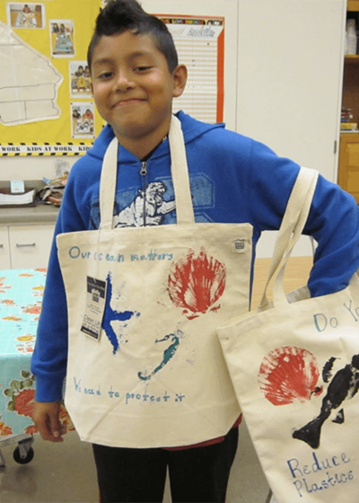 Young male student with dark hair and blue zip up sweatshirt with decorated tote bags hanging over his neck and off his shoulder in a colorful classroom setting.