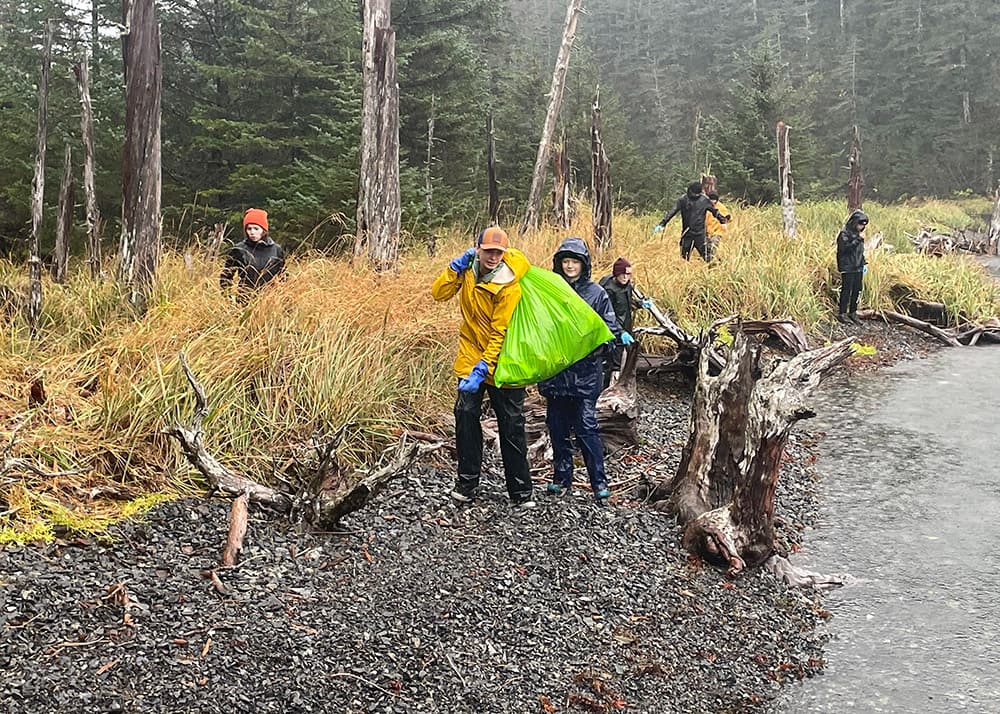 From left to right: Rocky beach with tall yellow and green grasses and trees and tree branches, student in black jacket and orange beanie, student in yellow jacket and blue gloves carrying green trash bag over his shoulder, five other students in the background, tree branches and waterway.