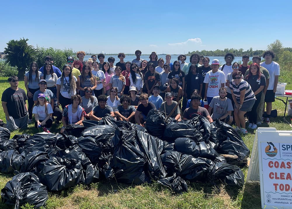 Large group of students and several adults with large black plastic trash bags on the grass with the ocean behind them. White fold up table and white foldable sign advertising cleanup on right.