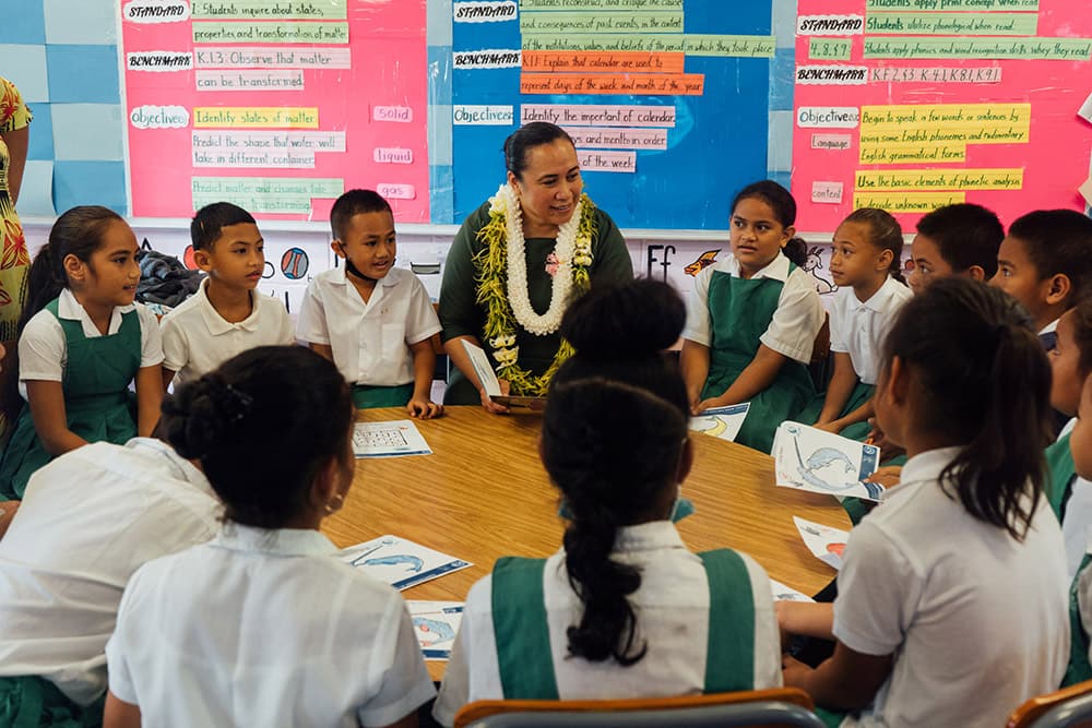 a woman wearing a necklace of flowers around her neck stands at a round table surrounded by young children dressed in school uniforms