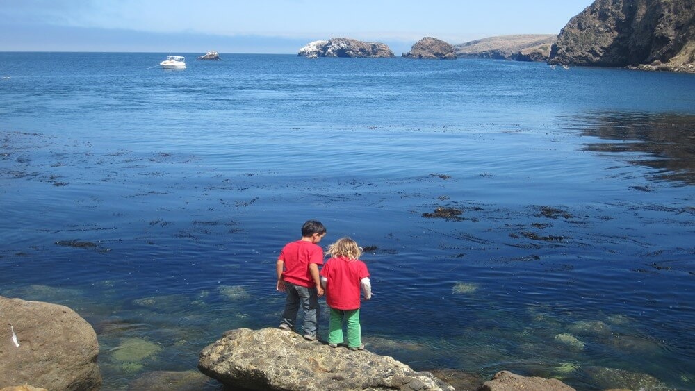 Two young children, one male with brown hair and one female with blonde hair, in red t-shirts on a rock looking into the blue water in the Channel Islands. In the background from left to right, there is a white boat, multiple large rocks sticking out of the water, two kayakers, and cliffs.
