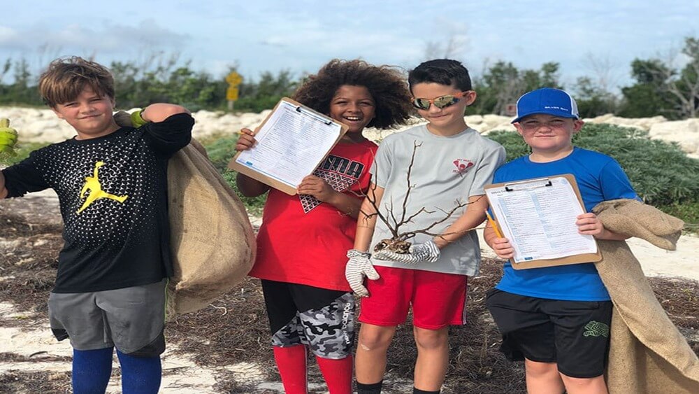 Four young boys, From left to right: boy with black shirt, gray shorts, and blue leggings with yellow gloves and filled burlap sack over his back. Boy smiling with a clipboard in a red tshirt, black camo shorts, and red leggings. Boy smiling with a gray tshirt, red shorts, sunglasses while holding a small tree with globes on. Boy smiling in blue hat and blue t shirt, black shorts, with clipboard and pencils and empty burlap sack in his arms.