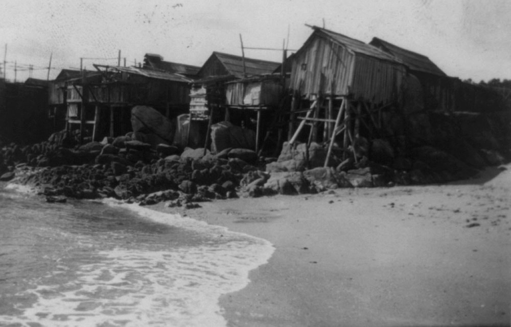 A historic photo shows wooden cabins built on sticks atop rocks near the water’s edge.