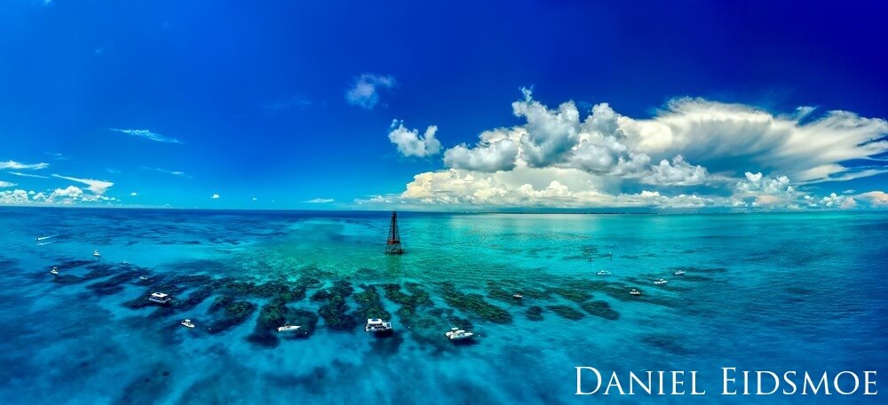 A panoranmic shot of several dive boats surrounding a lighthouse situated on a coral reef outrcropping.