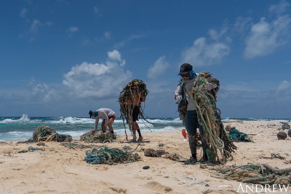 Marine debris technicians stand on a sandy beach picking up large amounts of nets, rope and trash. Waves crash into shore in the backgroundas the beach is littered with debris.