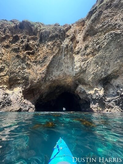 The edge of a kayak sits on clear water, and is pointed towards the entrance of a half-submerged rock cavern.
