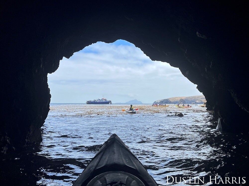 A group of kayakers is seen exiting from inside of a partially submerged cave.