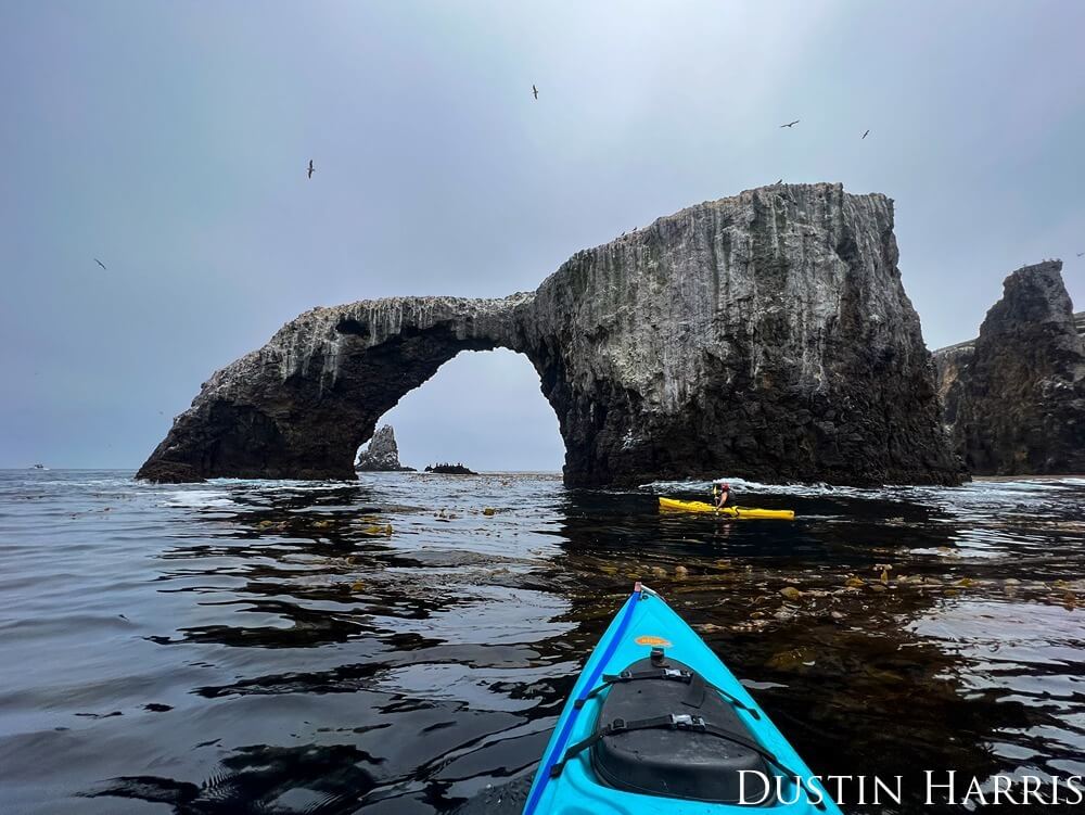 A group of kayakers are approaching a rock archway over the ocean.