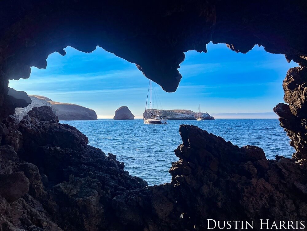 Sailboats are seen anchored beyond the mouth of a cave onshore.