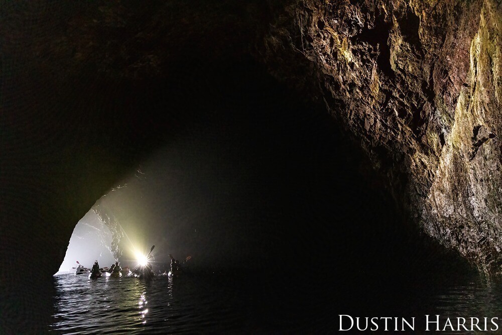 A group of kayakers exits a dark cave an paddles closer to the light coming from the mouth.