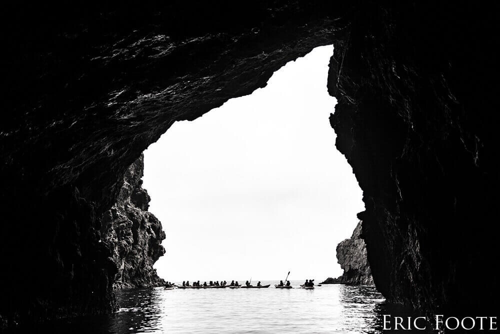 A group of kayakers is seen floating on the outside of a cave, pictured in black and white.