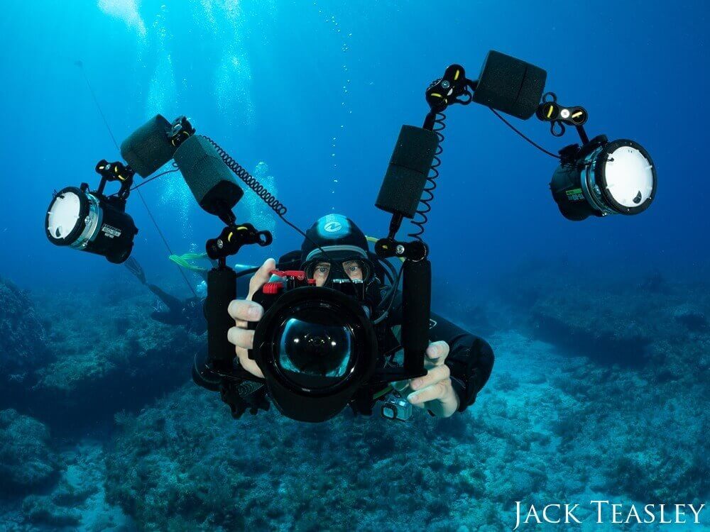 A diver is seen hovering holding a large camera, pointed at the photographer. 