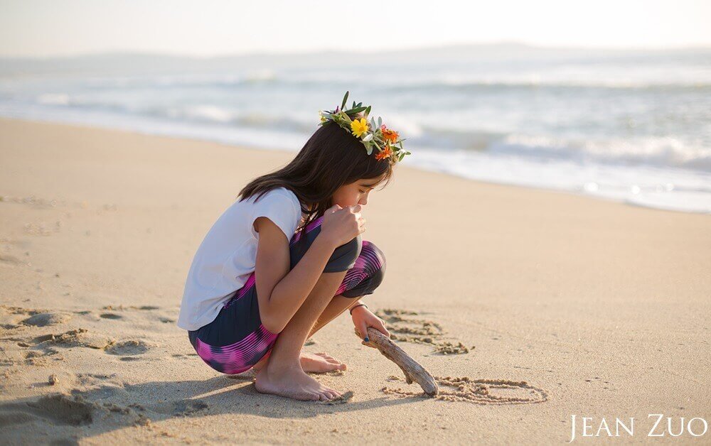A young girl wearing a flower crown is squatting on the beach writing in the sand with her finger.