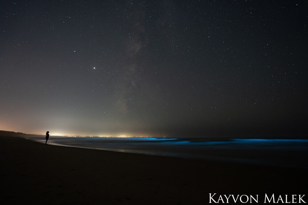 A silouette stands on the beach, looking up at the night sky illuminated by the glow of the city in the background