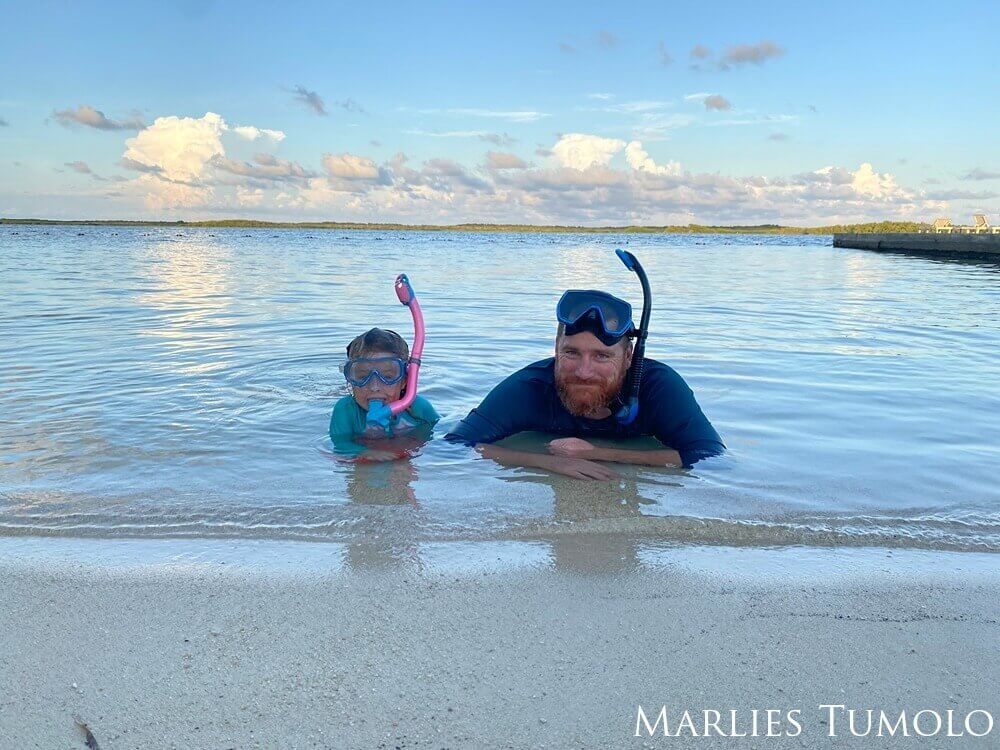 A father and daughter side by side in the water on the beach wearing snorkel gear on their heads.