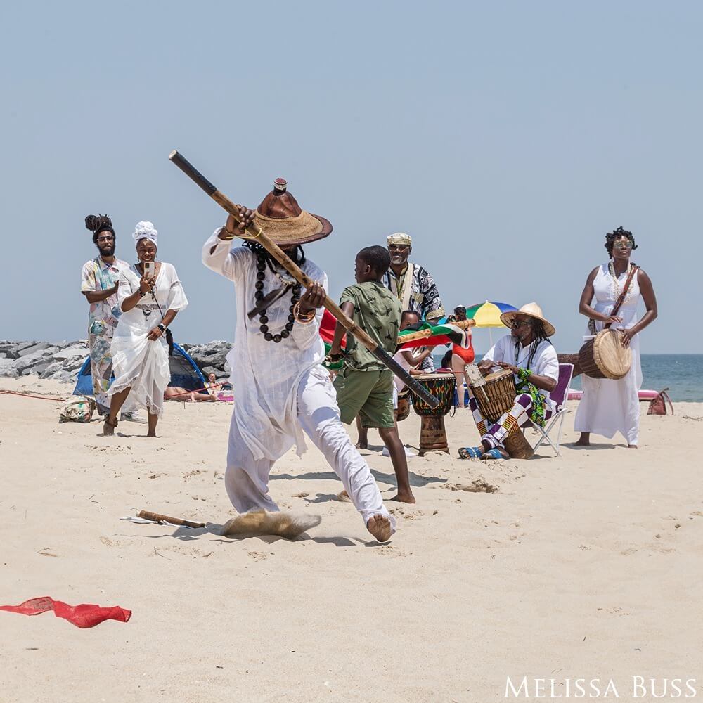 Men are seen dancing in the sand on the beach wearing ceremonial clothes as they particpate in the Sankofas Ceremony.