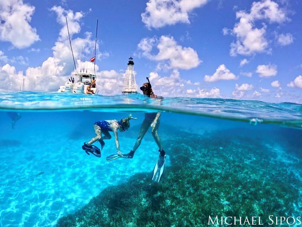 Two snorkelers are seen in the water in front of a boat and lighthouse surrounded by clear blue water.