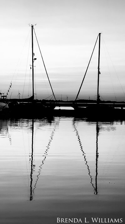 Black and white of two sailboats reflected in river.