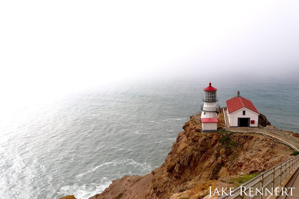 A narrow staircase on the right side leading down a brown rocky cliff towards the middle of the photo. At it's base a small white lighthouse and bunkhouse with red roofs. The surrounding area is a small visible portion of water mostly covered by white fog. 