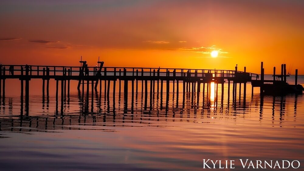 Colorful sunset over calm waters with pier and boat.