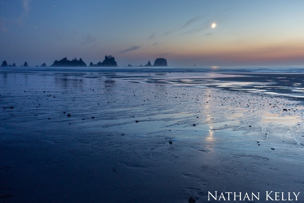 Moonlight and the twilight sky reflect on a beach etched with water rivulets after sunset.