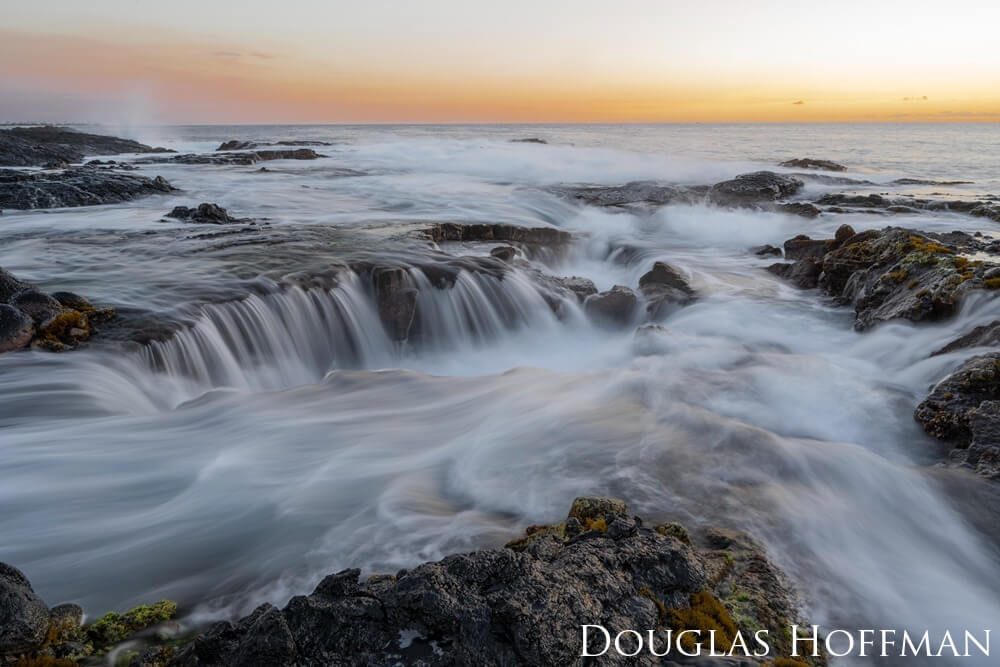 A colorful sunset behind the rocky coastline as waves crash over the rocks.