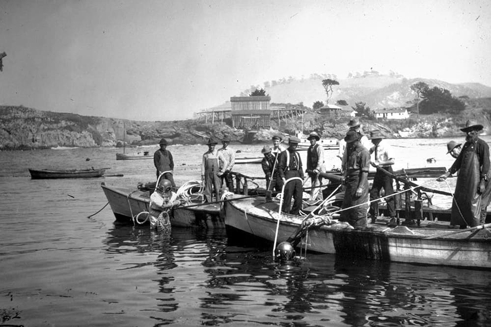 a scuba diver wearing a dive bell hangs onto the side of a small boat with several men standing in it, next to another boat with more people in it