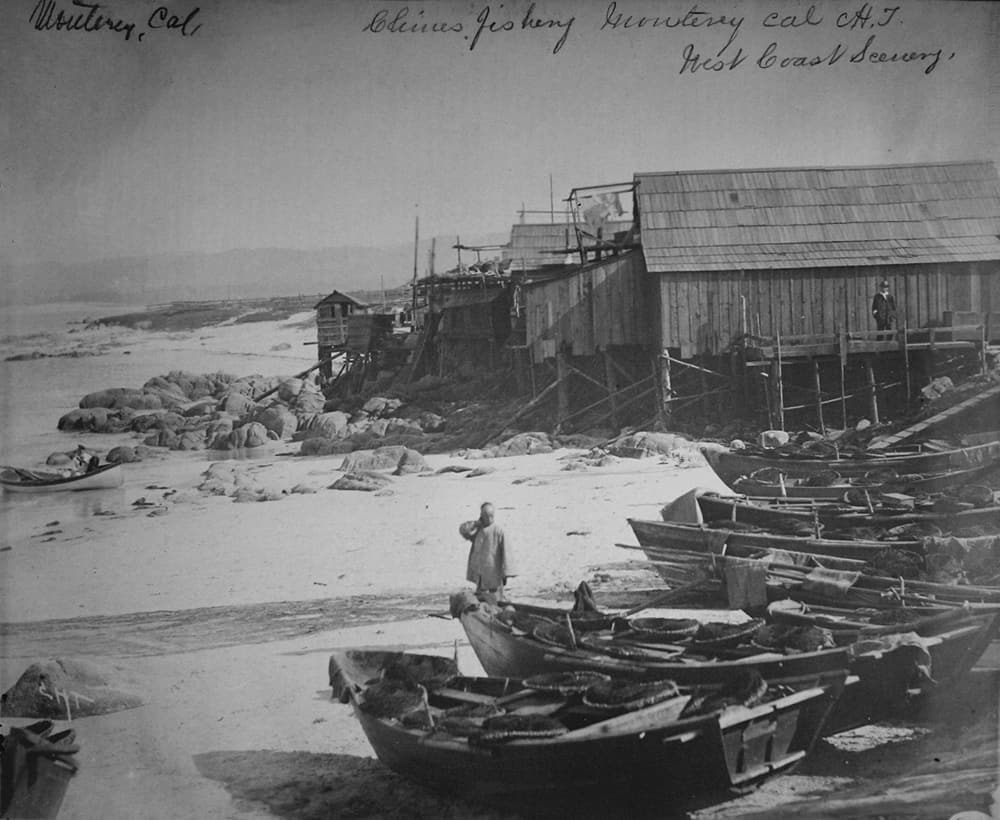 A historic photo shows a row of small wooden boats pulled up on shore beside a row of wooden cabins built on sticks on top of rocks