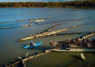 A person in a blue kayak paddles between two shipwrecks at the surface of the water, with additional shipwreck pieces and land visible in the background.