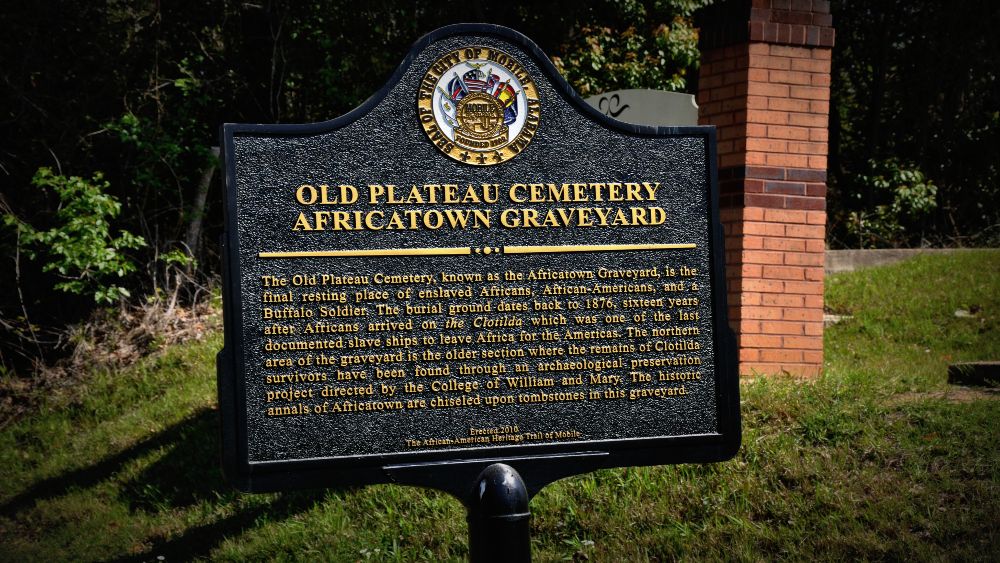 A black metal sign with yellow text that reads old plateau cemetery africatown graveyard
