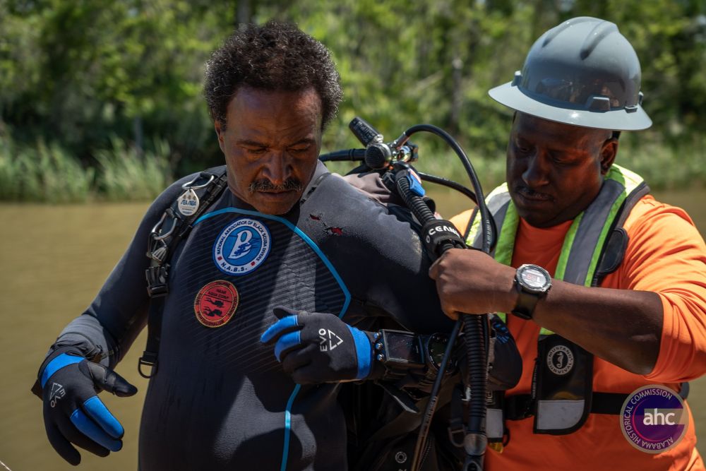 a person wearing a hard hat helps another person don their scuba gear