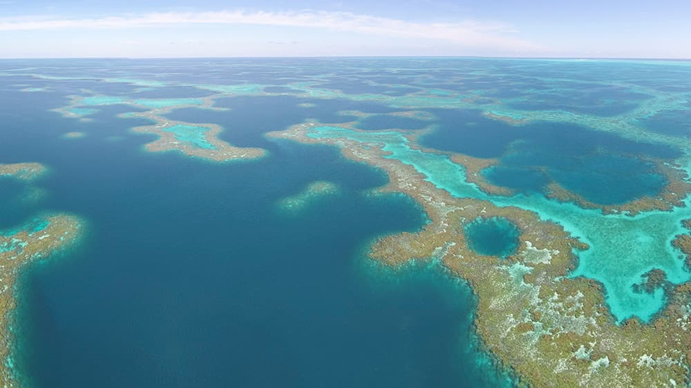 aerial photo of teal water with pockets of lighter turquoise waters surrounded by orange, brown, and yellow, coral reefs that rise up to the ocean surface
