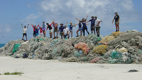 15 volunteers standing on a large stack of multicolored derelict fishing nets on sandy beach with blue skies and clouds above.