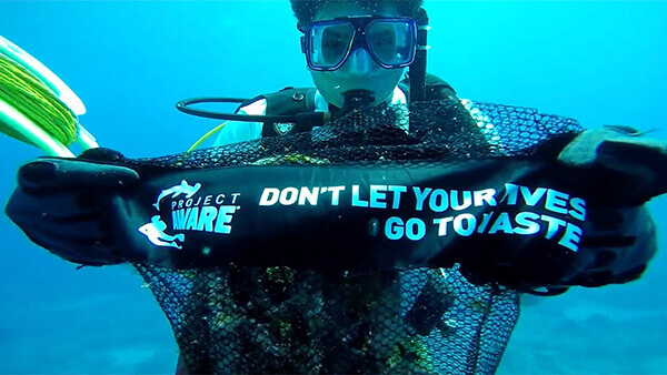 Diver underwater with black netted bag with white writing “Project AWARE: DON’T LET YOUR DIVES GO TO WASTE””