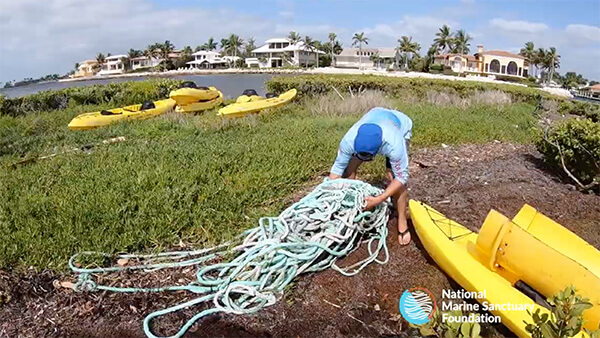Volunteer in blue long sleeve and blue hat moving green discarded nets on dirt path with grass behind and yellow kayaks stacked by waterfront.