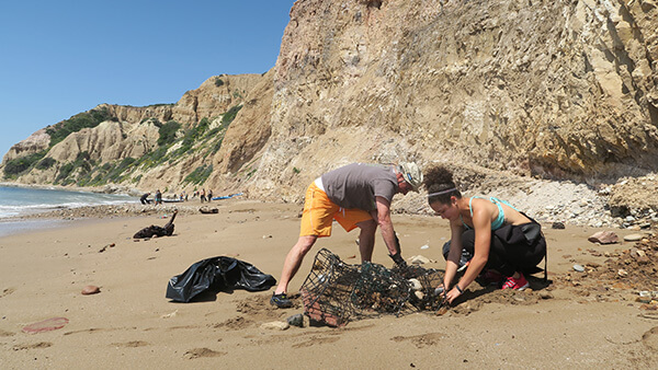 Two volunteers removing lost fishing gear on a beach with cliffs overhead and other volunteers in the distance.
