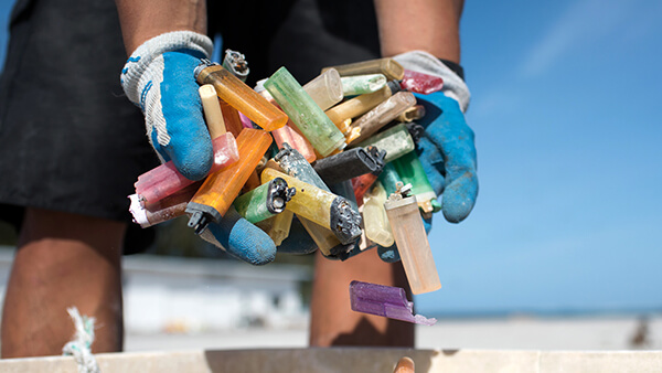 Multicolored plastic lighters being dropped into a bin after a beach cleanup by a volunteer with blue and white gloves and black athletic shorts.