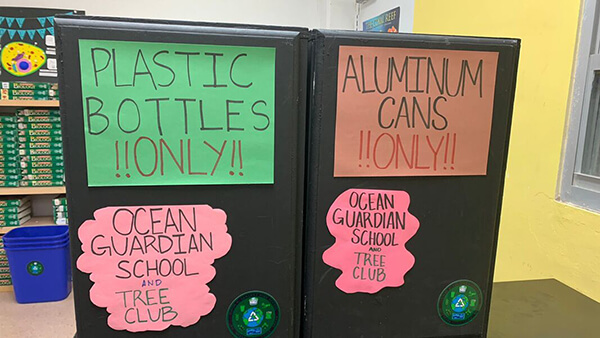 Recycling bins at Ocean Guardian School with green sign reading “PLASTIC BOTTLES !!ONLY!!” and an orange sign reading “ALUMINUM CANS !!ONLY!!” on two black recycling containers. 