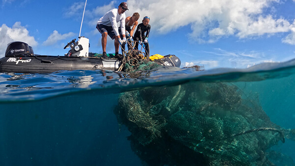 Three individuals pulling large bunch of entangled nets out of the ocean onto boat.