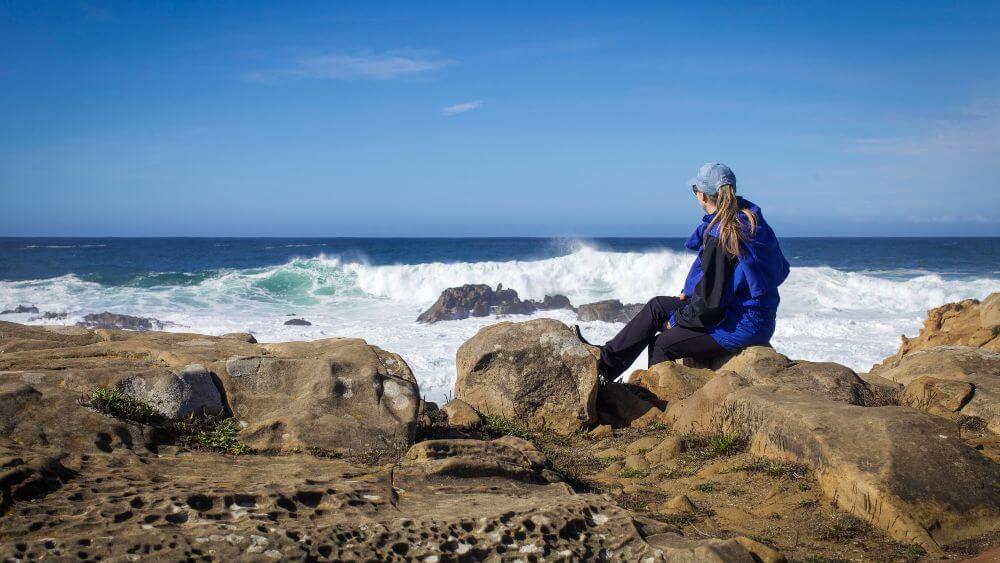 A woman in a blue jacket sits on a rock facing the ocean waves in the distance in NOAA’s Greater Farallones National Marine Sanctuary.