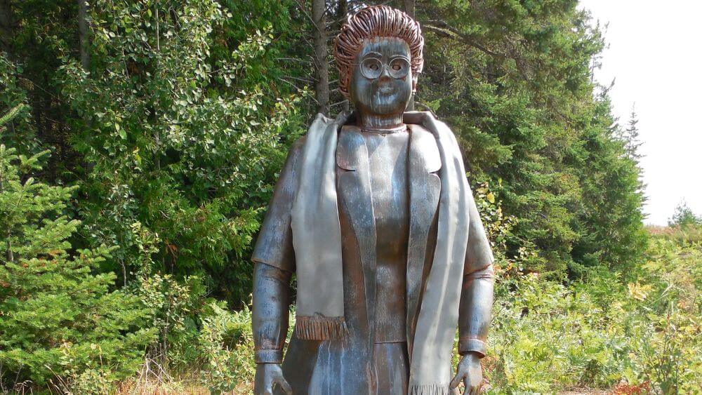 a metallic, lifesize statue of Anna Garrity stands against a backdrop of trees