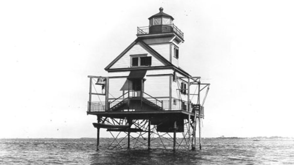 a lighthouse and keeper’s house sit in stilts offshore of a low sandy island