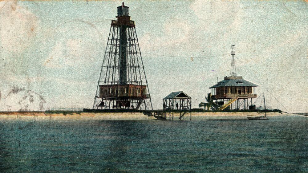 a lighthouse with an open metal frame and a lightkeeper’s house on stilts sit on a low sandy island